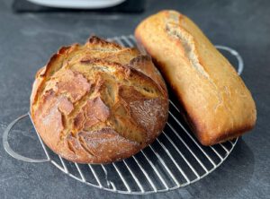 Ein rundes und ein kastenförmiges Brot mit drunkler Kruste auf einem Kuchengitter.